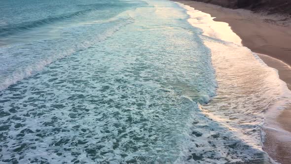 Aerial view of waves crash to shore at sunset alt