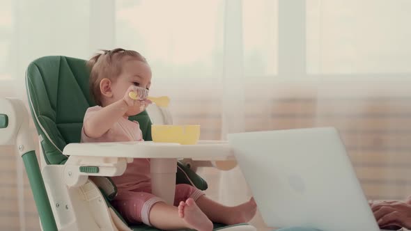 Child Eats Breakfast Porridge While Sitting at a Table Mom Uses a Laptop alt