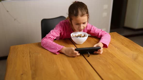 Little girl eating cereal with milk and play game on smartphone