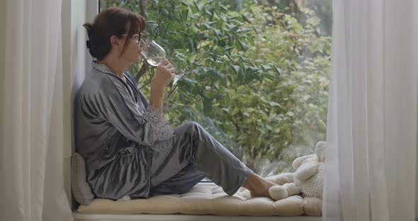 Wide Shot of Carefree Caucasian Female Retiree Sitting on Windowsill Drinking Wine From Glass alt