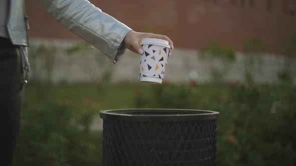 A Woman Throws a Drunk Cardboard Cup with Coffee Into a Trash Can in a Park