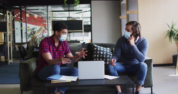 Diverse business people wearing masks using laptop and goign through paperwork in modern office alt