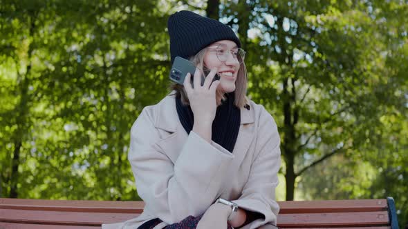 Young Girl Sitting on a Park Bench in Autumn Talking on the Phone and Smiling