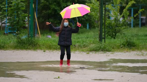 Little girl with an umbrella in a medical mask and rubber boots jumps through puddles