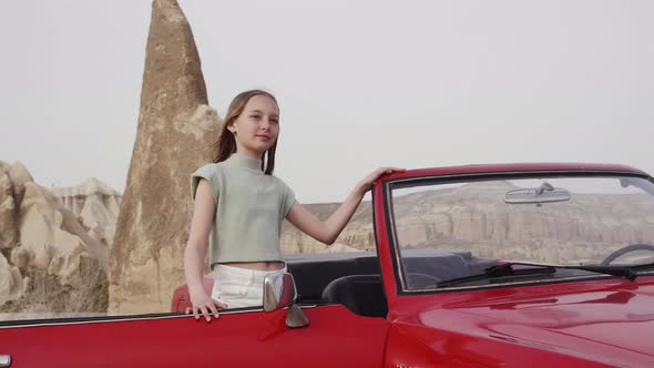 Pretty Young Girl Stand By Open Door of Red Retro Cabrio Car Against Rock Formations Mountains and