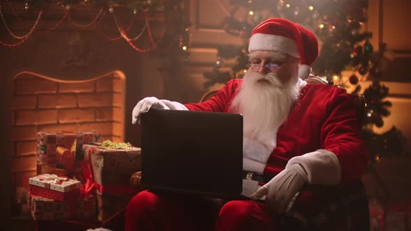 Modern Santa Claus. Cheerful Santa Claus Working on Laptop and Smiling While Sitting at His Chair alt