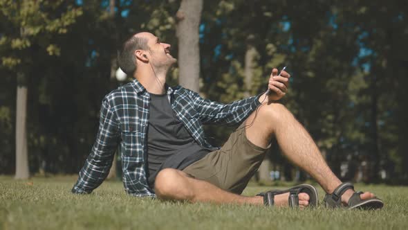 Young Handsome Guy in a Plaid Shirt Listens to Music While Sitting on the Grass in the Park alt