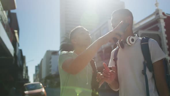 Two happy mixed race male friends standing, talking and using smartphone in the street alt