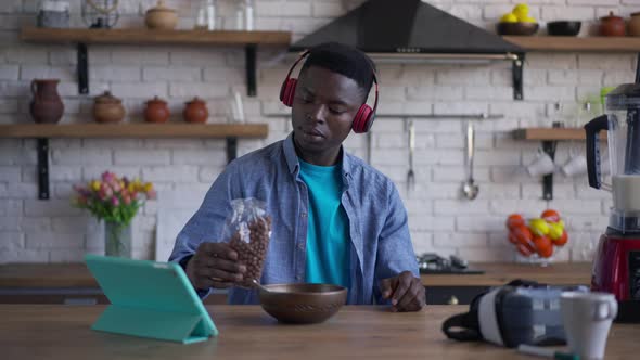 Portrait of Young African American Man Pouring Breakfast Cocoa Balls in Bowl Sitting in Kitchen at alt