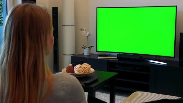 A Blonde Woman Sits on a Couch in a Cozy Living Room and Watches a TV with a Green Screen alt