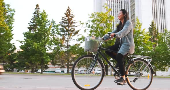 Asian girl is riding bicycle in city, carrying bag from supermarket in basket. Young woman