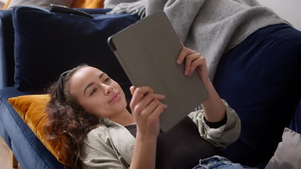 Overhead Shot Of Young Woman Relaxing At Home Lying On Sofa Reading Book On Digital Tablet alt