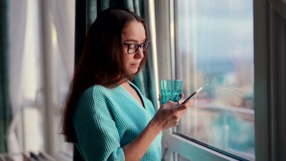Nervous and Excited Woman is Typing Message By Mobile Phone Standing with Glass Near Window in Room alt