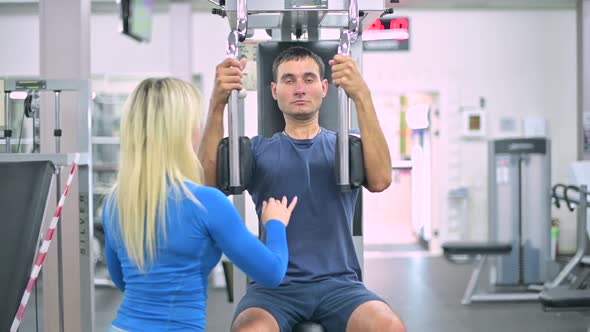 skinny guy does an exercise on an machine under the supervision of a trainer alt