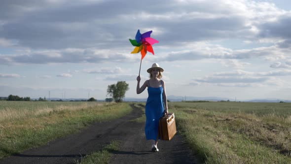 Girl in blue dress with suitcase and pinwheel on country road in summer. alt