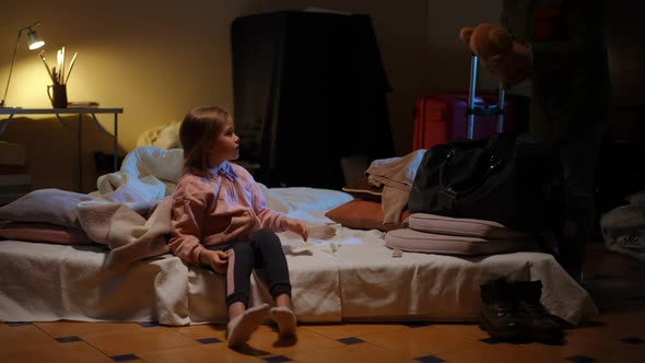 Charming Cute Little Girl Passing Toy to Woman Packing Suitcase in Basement Floor at Home alt