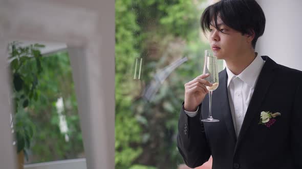 Portrait of Serious Young Asian Man in Wedding Suit Looking Out the Window Drinking Champagne alt