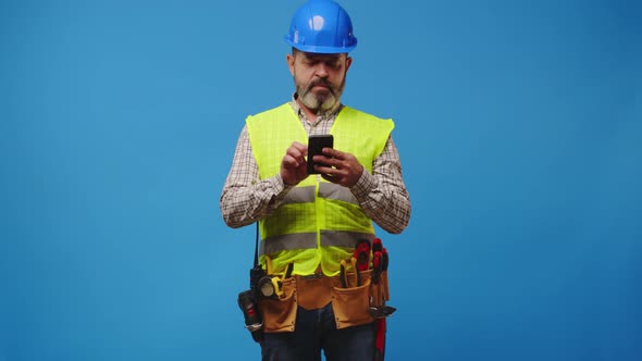 Senior Man Builder in Vest and Hardhat Using His Smartphone Against Blue Background