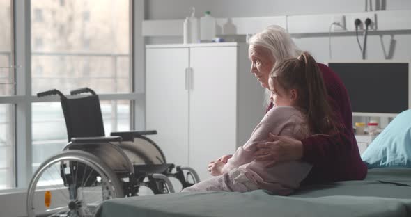 Senior Woman and Little Girl Sitting on Hospital Bed and Hugging alt