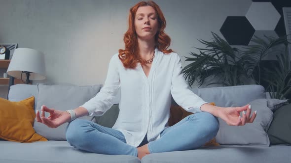 Red Haired Woman Making Mudra Gesture Sitting in Lotus Position on Couch at Home alt