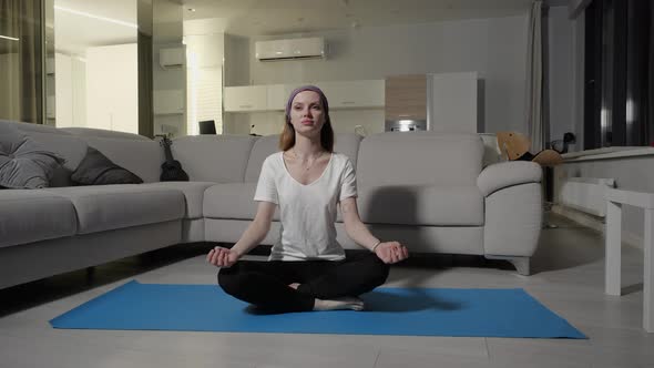 A Young Girl in a White Tshirt Sits in a Lotus Pose on a Blue Yoga Mat at Home