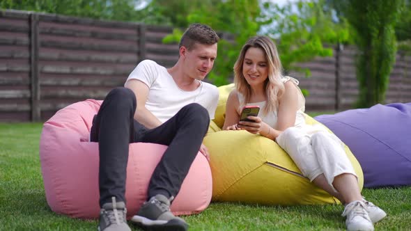 Wide Shot Positive Millennial Couple Sitting on Bag Chairs on Green Meadow Scrolling Social Media on alt