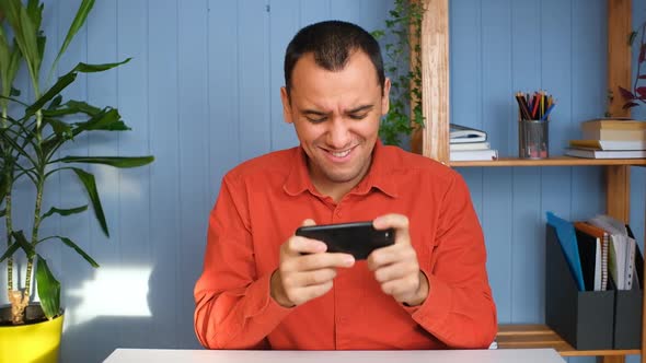 Happy Man Is Playing Video Game at Home Office Using Modern Smartphone
