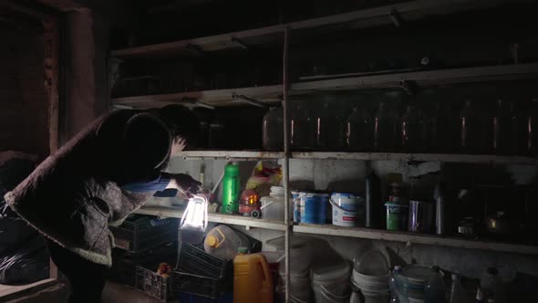 An Elderly Woman Looking for Canning on the Shelves in a Dark Old Cellar Using a Lantern