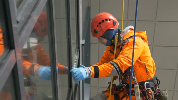 Industrial Climber in Orange Overalls and Helmet Washes Windows on Tall Building Outside. alt