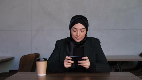 Young Muslim Woman in Hijab Playing Mobile Game in Cyberspace at Cafe Whit Coffee