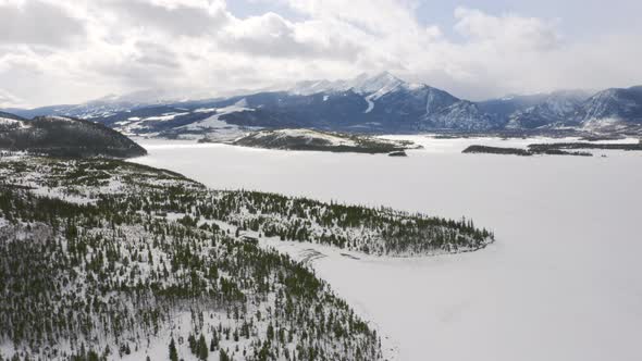 Aerial moving forward to reveal icy, frozen lake surrounded by snow and green pine trees with large alt
