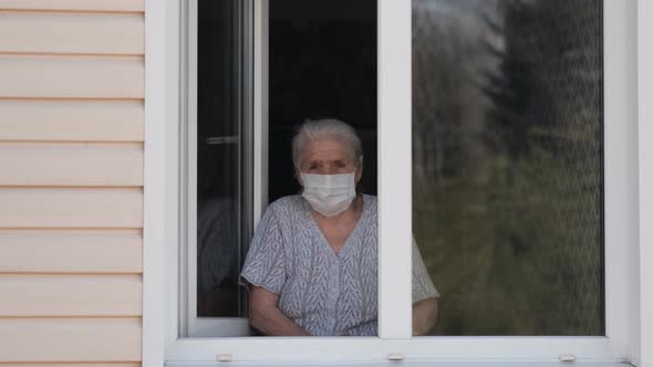 Sad Senior Woman in a Medical Mask Stands at Home at the Window and Look at the Street.