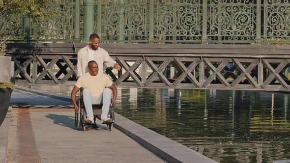 Young Loving Black Guy Accompanies Aged Disabled Person Sitting in Wheelchair During Walk alt