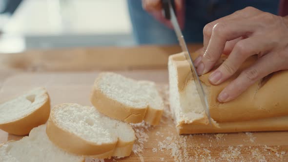 Hands of woman chef holding knife cutting whole grain bread on wooden board on kitchen table. alt