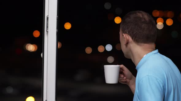 Man drinks tea and looks at the lights of the night city, back view alt