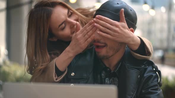 Absorbed Gay Man Surfing Internet on Laptop As Woman Closing Eyes with Hands Entering Sidewalk Cafe alt