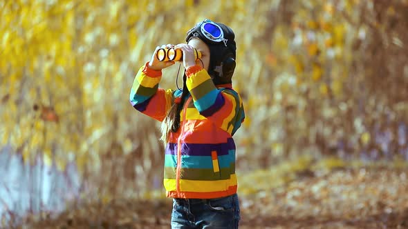 Cute little girl in a pilot's helmet and glasses looking through binoculars in the autumn Park