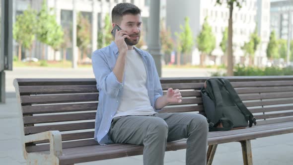 Angry Young Man Talking on Phone While Sitting on Bench alt