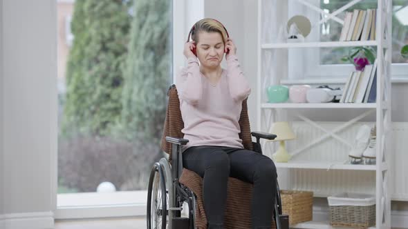 Portrait of Relaxed Joyful Disabled Woman in Wheelchair Putting on Headphones and Moving To Music alt