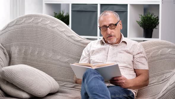 Focused Aged 70s Grandfather Reading Book Sitting on Couch alt
