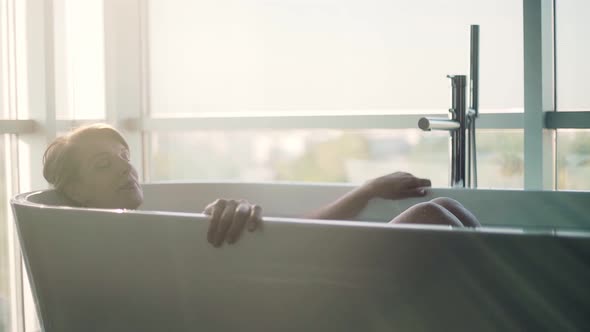 Retired woman taking bath in bathtub against window alt