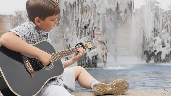 A Guy in a Shirt and Shorts Plays the Guitar Close to the Water Fountain in the Park alt