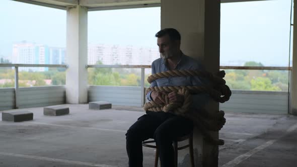 Businessman in Shirt Sit Tied with Thick Rope in Parking Lot alt