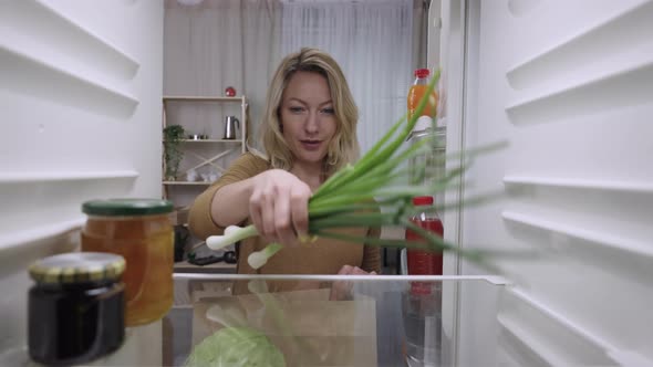 Young Woman Opens Refrigerator Door Holding Paper Bag of Groceries and Putting Food in Fridge alt