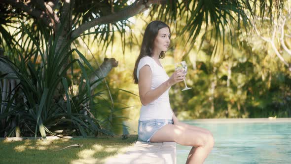 Woman Sits on Edge of Pool Splashes Water with Feet and Drinks Water From Glass alt