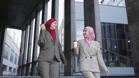 Two Young Muslim Women Wearing Hijab Headscarf Look at Phone, Walking Together Down Stairs Drinking