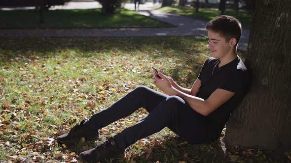 Boy Is Sitting Under a Tree with a Smartphone alt