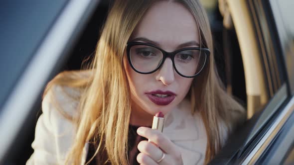 Pretty Woman Looking in Rear View Mirror and Applying Red Lipstick in Car alt