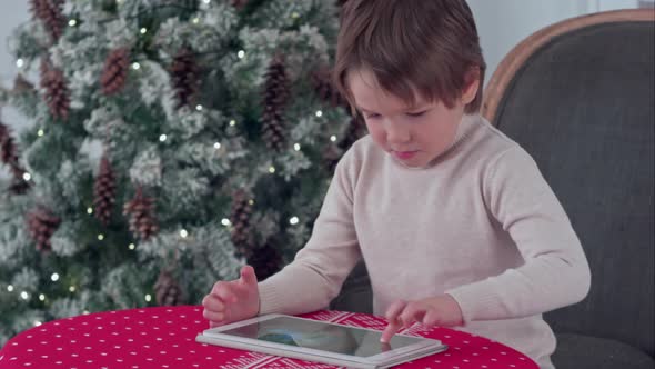 Boy Playing Games on His Tablet Sitting at the Table Near the Christmas Tree alt
