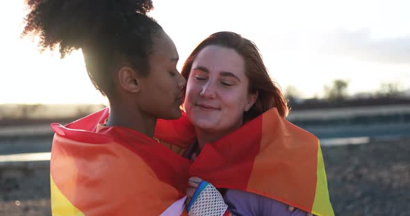 Young lesbian couple having tender moment at sunrise wearing LGBT rainbow flag alt